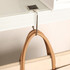A square silver desk hook holding a beige bag, mounted on a white surface.