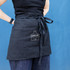 A black waist apron with a front pocket and a tied waist, held by a person against a blue background.