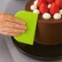 A bright green plastic flexible bowl scraper being used to smooth icing on a chocolate cake topped with strawberries and cream.