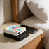 A square power bank in white with a floral design sits atop a black power bank on a bedside table next to a smartphone.