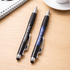 A pair of pens, one black and one blue, with grips, resting on a wooden surface near a cup and notebook.
