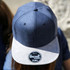 A navy blue cap with a grey brim and a prominent logo on the front.