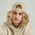 A beige cap with a flat peak is worn by a model with curly blonde hair, featuring a rear adjuster.
