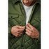 A man in a green quilted jacket adjusts the zipper, revealing a grey top underneath. The jacket features snap buttons and a logo.