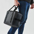 A cooler bag in black with sturdy handles, being carried by a person wearing jeans. Features a logo.