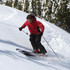 A skier wearing a red jacket and black pants navigates through snow-covered terrain with ski poles.