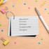 Pearlescent index cards in white, bound by a ring, alongside a pen and colourful paper clips on a wooden surface.