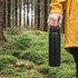 A drink bottle in matte black, featuring a handle, held by a person in a yellow jacket amidst a forested background.