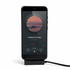 A black phone on a stand displaying a music player interface with a charging cable attached.