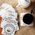 A set of square cardboard coasters in white, featuring a logo, next to a black coffee cup on a textured surface.