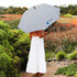 A grey umbrella with a logo is held by a person in a white dress, surrounded by garden scenery.