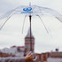 A transparent eight-panel umbrella with a logo, held up against a blurred city background.