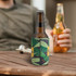 A drink bottle is inside a green reversible foldable stubby holder with a leaf design, placed on a wooden table.