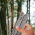 A silver survival multi-tool is held in a hand, set against a background of blurred bamboo. It has a logo on it.