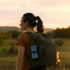 A woman with a ponytail wearing a brown top and carrying a green backpack, set against a sunset landscape.