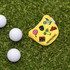 A yellow custom mallet putter cover with various patches, placed on grass near three white golf balls.