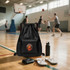 A black multi-pocket drawstring backpack sits on a gym floor beside a drink bottle and flip-flops, with players in the background.