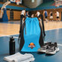 A blue and black sports drawstring backpack sits on a gym floor, beside a drink bottle, towel, and shoes.