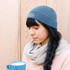 A blue round beanie worn by a woman with long braids, sitting and holding a blue cup. The beanie features a logo.