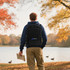 A black expandable laptop backpack is worn by a person standing by a lake, with autumn foliage in the background.
