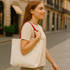 A woman with long hair carries a beige 340gsm canvas tote bag with red handles in an outdoor setting.