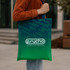 A person holding a green and blue gradient tote bag with a logo. The bag features a sturdy design.