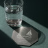 An octagonal metal coaster with a silver finish rests on a green surface beside a glass of water.