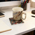 A ceramic coaster with a colourful design holds a patterned mug on a desk, surrounded by stationery and a lamp.