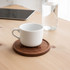 A round wooden coaster with a natural finish holding a white ceramic cup on a wooden table.