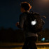 A reflective heart-shaped keychain attached to a backpack, glowing in the dark, with a person walking in the background.