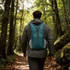 A teal large packable camping backpack is worn by a person walking on a trail through a lush, green forest.