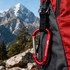 A red carabiner keychain attached to a black and red backpack, set against a mountainous backdrop.