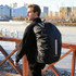 A black reflective backpack rain cover is worn by a person outdoors near a water body and buildings. It has a logo.