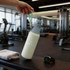 A drink bottle in pale green and grey, featuring a logo, placed near a towel and a watch in a gym setting.