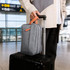 A grey classic toiletry bag with a tan handle sits atop a black suitcase, at an airport terminal.