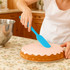 A blue spatula is being used to spread icing on a cake, set on a floral tablecloth with a mixing bowl nearby.