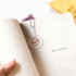A transparent PVC hand fan with a keychain, featuring a circular shape and a branding logo, rests on a book page.