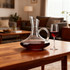 A glass wine decanter with a handle and deep red wine, beside a filled wine glass, on a wooden table.