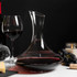 A glass wine decanter with a curved neck, filled with red wine, beside a wine glass, cheese, and grapes.