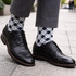 A pair of black leather shoes with grey and black checked socks, set against a stone pavement backdrop.