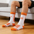Sport socks with a white base and orange detailing, featuring a logo, displayed on a person's feet while seated.