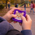 A purple square fidget keychain with a cat design, held in hands outdoors near a playground.