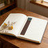 A brown PU bookmark with a teal accent rests on an open notebook beside dried flowers on a wooden table.