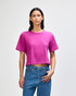 A woman wears a bright pink boxy T-shirt with short sleeves, paired with blue jeans. The shirt has a logo.