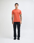 A model wearing an orange unisex short sleeve t-shirt and beige pants, standing casually. The t-shirt has a logo.