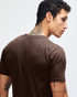 A brown short sleeve T-shirt with a fitted design, featuring a logo on the back. The model has tattoos visible.