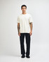 A unisex heavyweight cotton T-shirt in cream, paired with blue cargo pants and sneakers. The model has curly hair and a neutral pose.