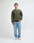 A unisex crewneck sweatshirt in olive green, paired with black cargo pants, worn by a model. Features a logo.