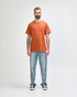 An adult wearing an orange Gildan Heavy Cotton T-shirt, paired with black pants and black shoes. The shirt has a logo.