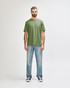 A man stands wearing an olive green Gildan Softstyle adult T-shirt paired with light blue jeans.